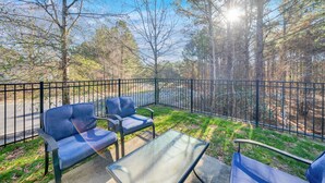 An outdoor seating area with two blue chairs and a glass table, surrounded by a black metal fence, overlooking a road and trees. - Stylish Luxe One-Level Twnh Steps from Neuse Trail (Raleigh)