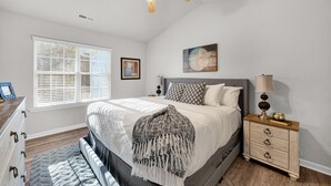 A bedroom with a bed, a nightstand, a ceiling fan, a window, and a painting on the wall. - Stylish Luxe One-Level Twnh Steps from Neuse Trail (Raleigh)