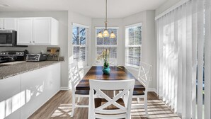 A modern kitchen with white cabinets, a wooden dining table, and a chandelier. - Stylish Luxe One-Level Twnh Steps from Neuse Trail (Raleigh)