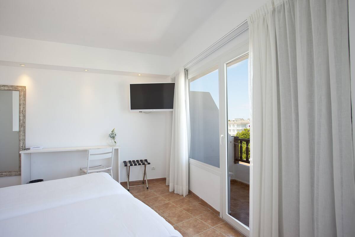 A hotel room with a bed, a desk, a chair, a TV mounted on the wall, and a balcony with curtains.