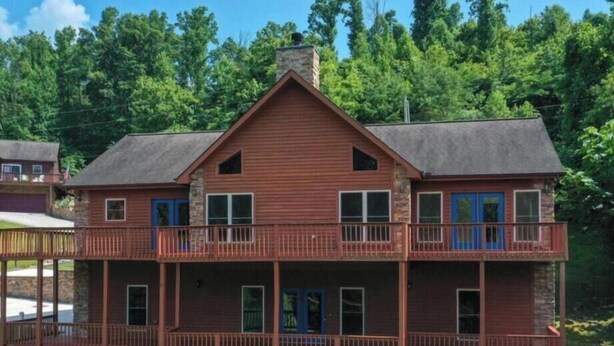 A two-story wooden house with a balcony and a stone chimney, surrounded by greenery.