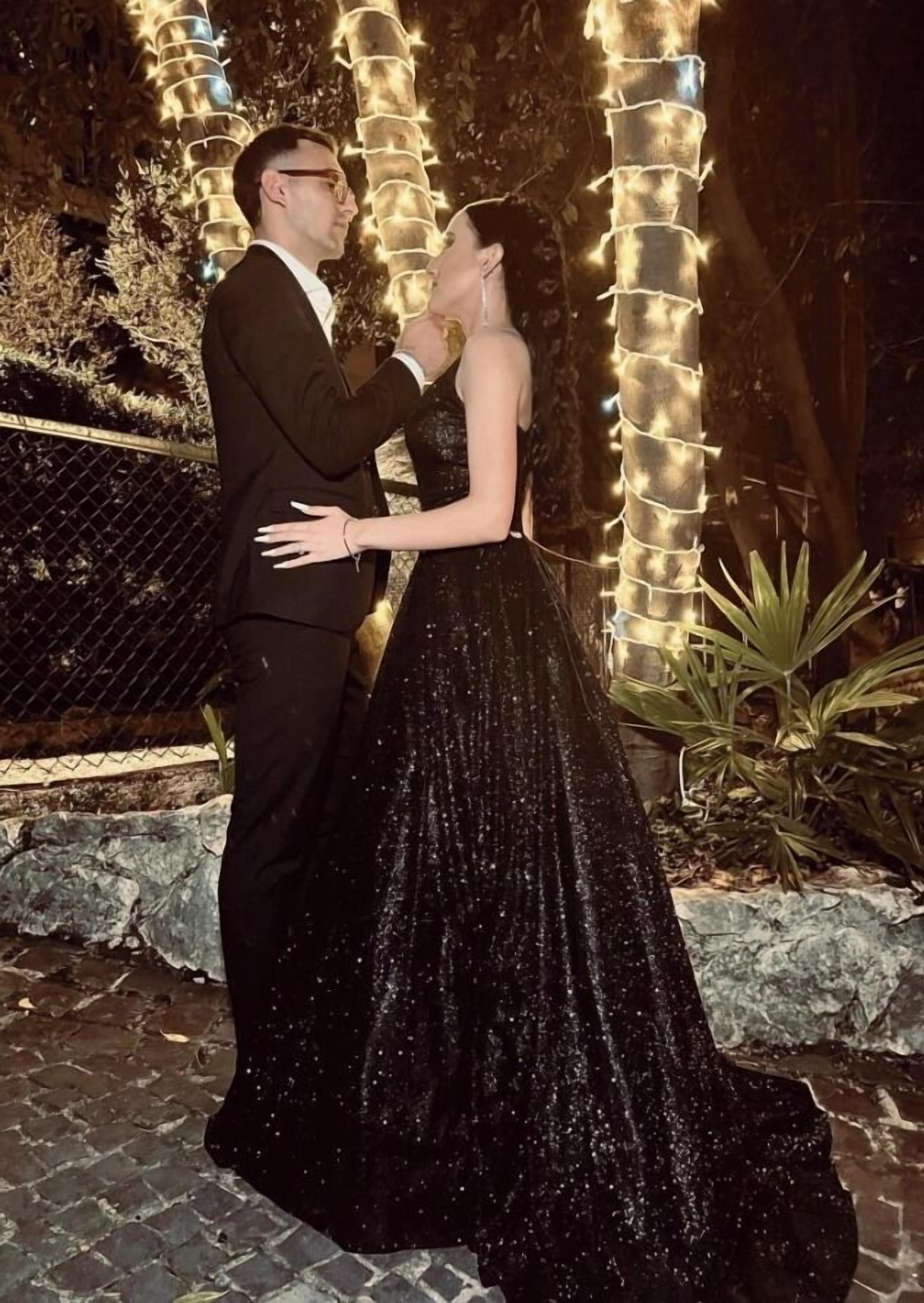 A couple embracing in a formal setting with decorative lights and a stone wall in the background.