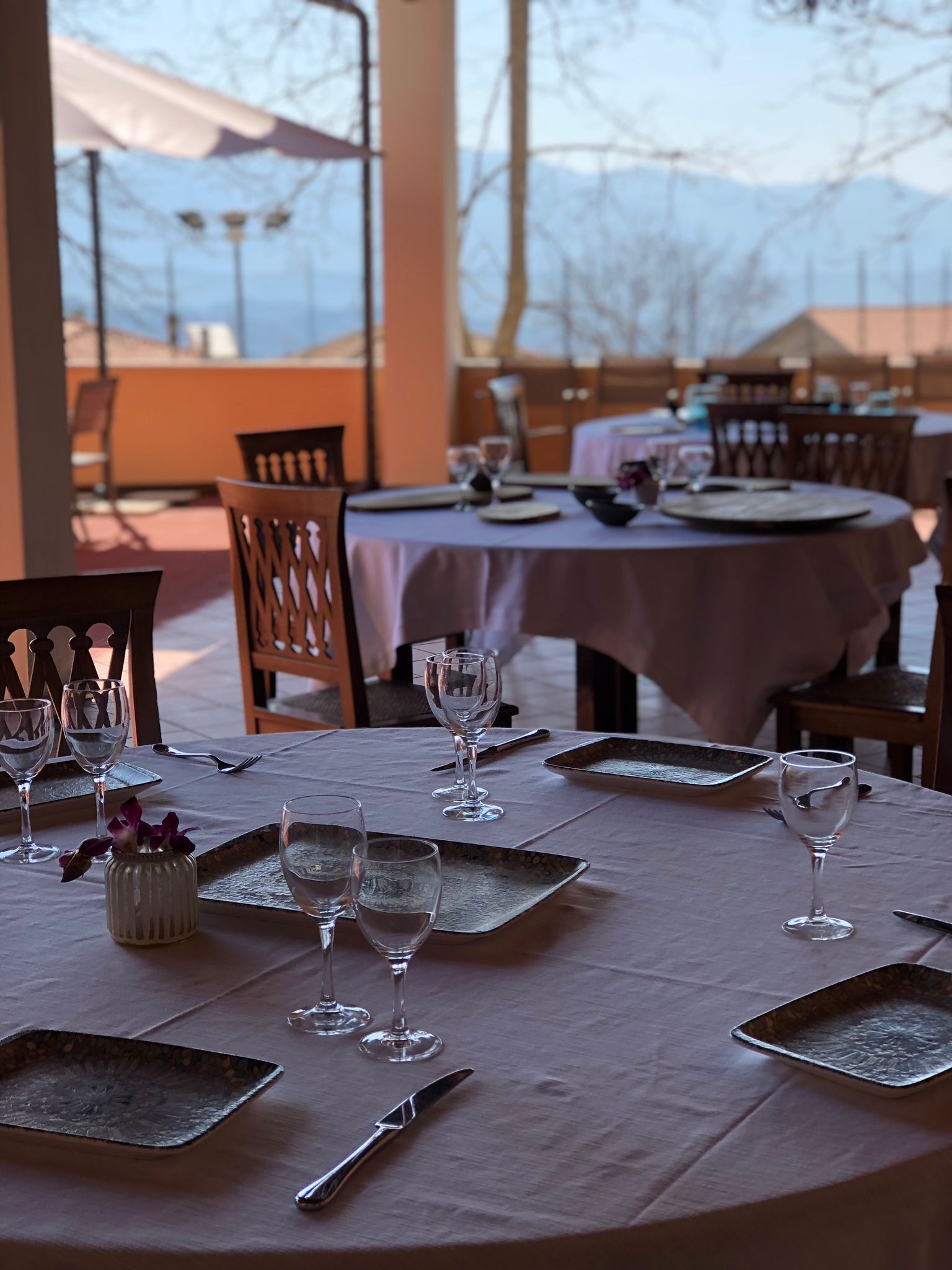 Uma área de refeições com mesas redondas preparadas para uma refeição, com taças de vinho vazias, pratos e talheres.