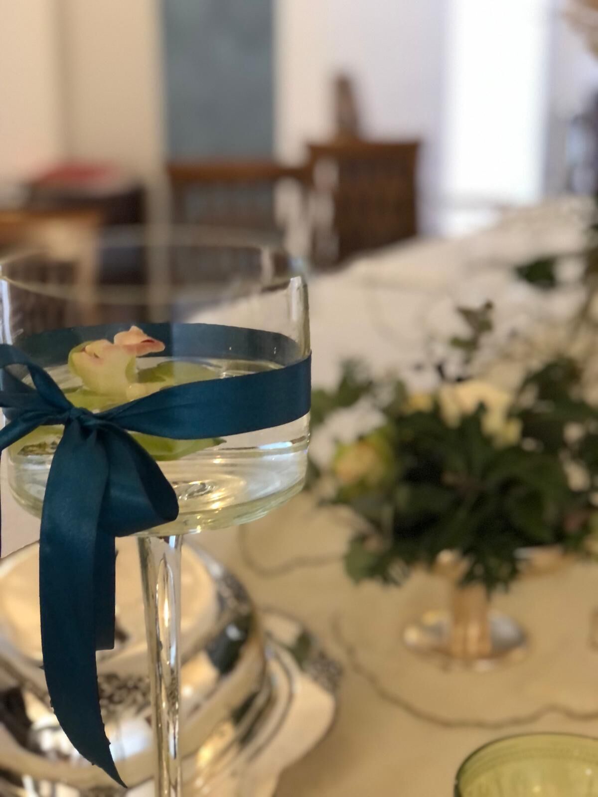 Um copo com uma fita azul e uma flor, colocado sobre uma mesa com um arranjo floral ao fundo.