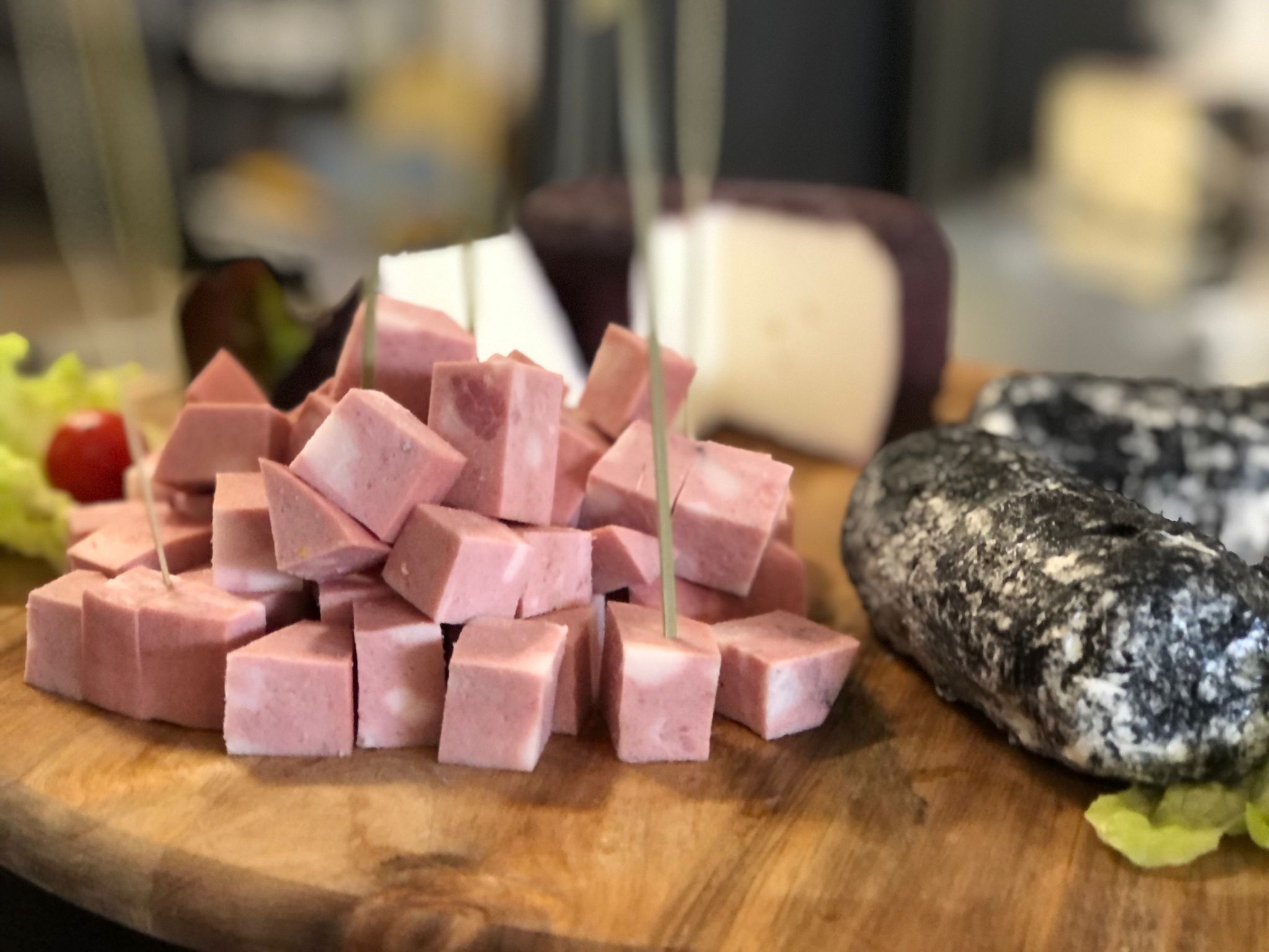A wooden cutting board with cubed pink ham, a whole cheese, and a sliced black and white cheese.