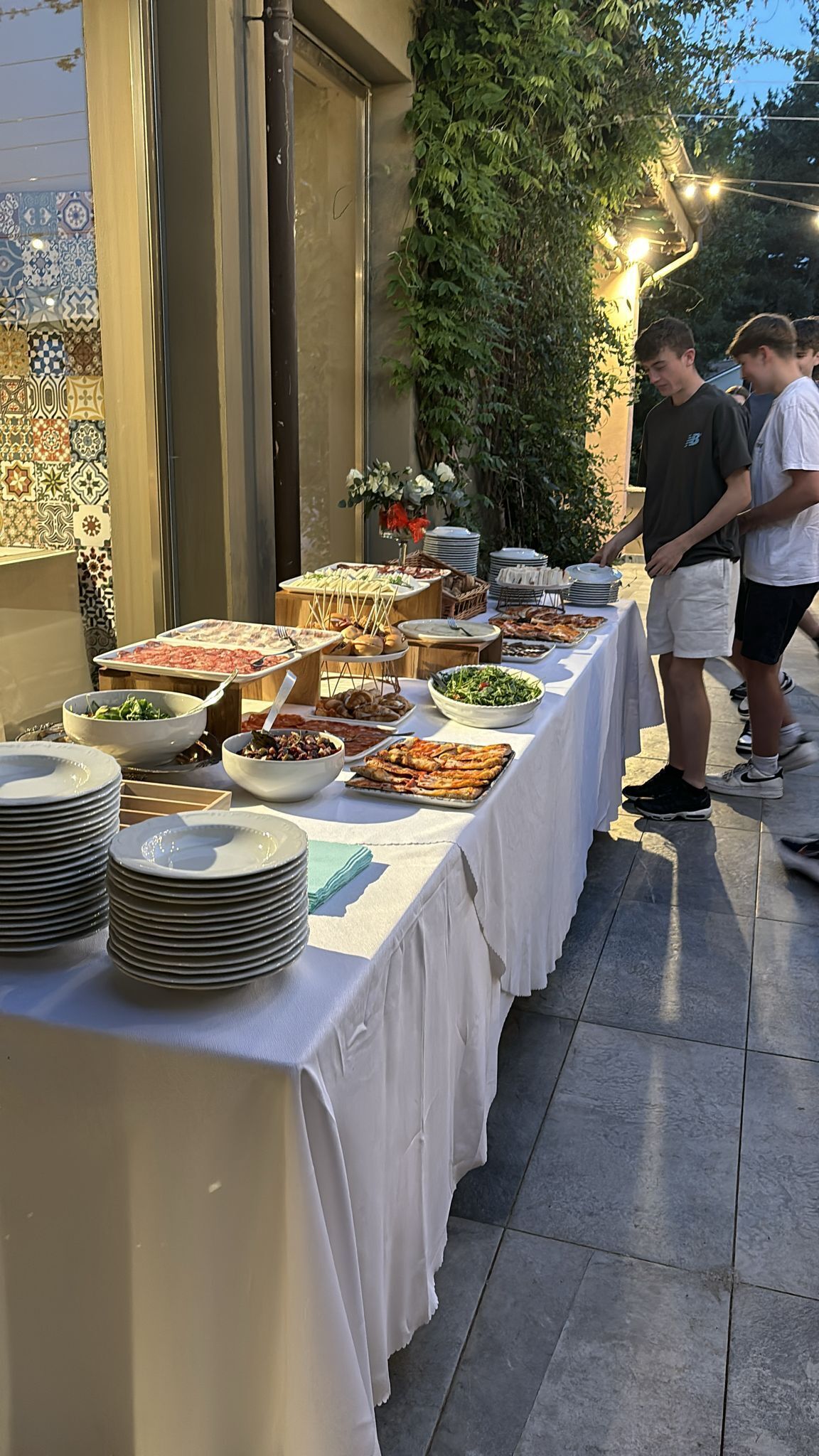 Montaje de bufé al aire libre con diferentes alimentos y platos, piso de baldosas y pared con un diseño decorativo de azulejos.