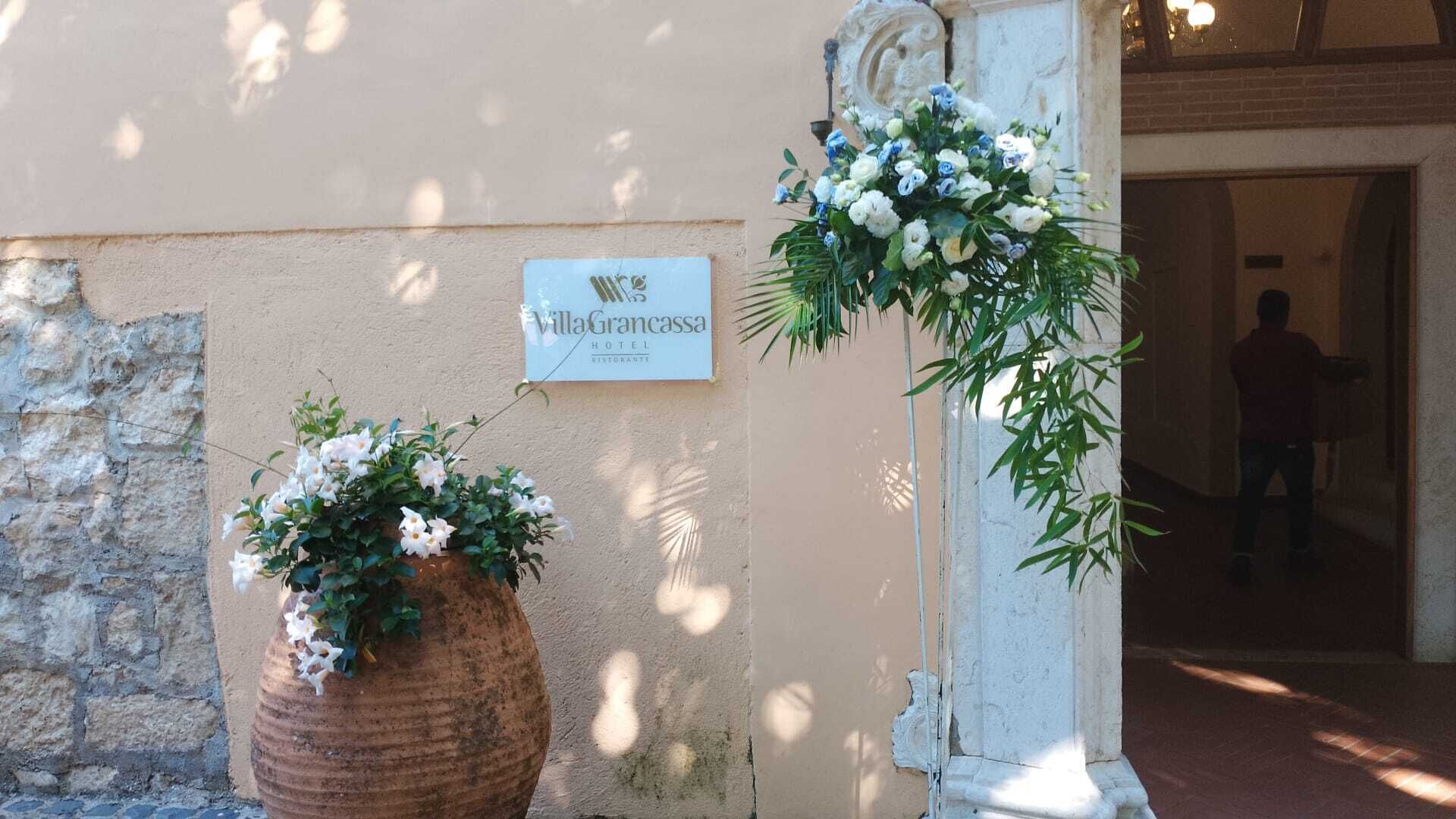 Señal del Hotel y Restaurante Villa Gracassa, una planta en maceta con flores blancas y un acceso con una persona al fondo.