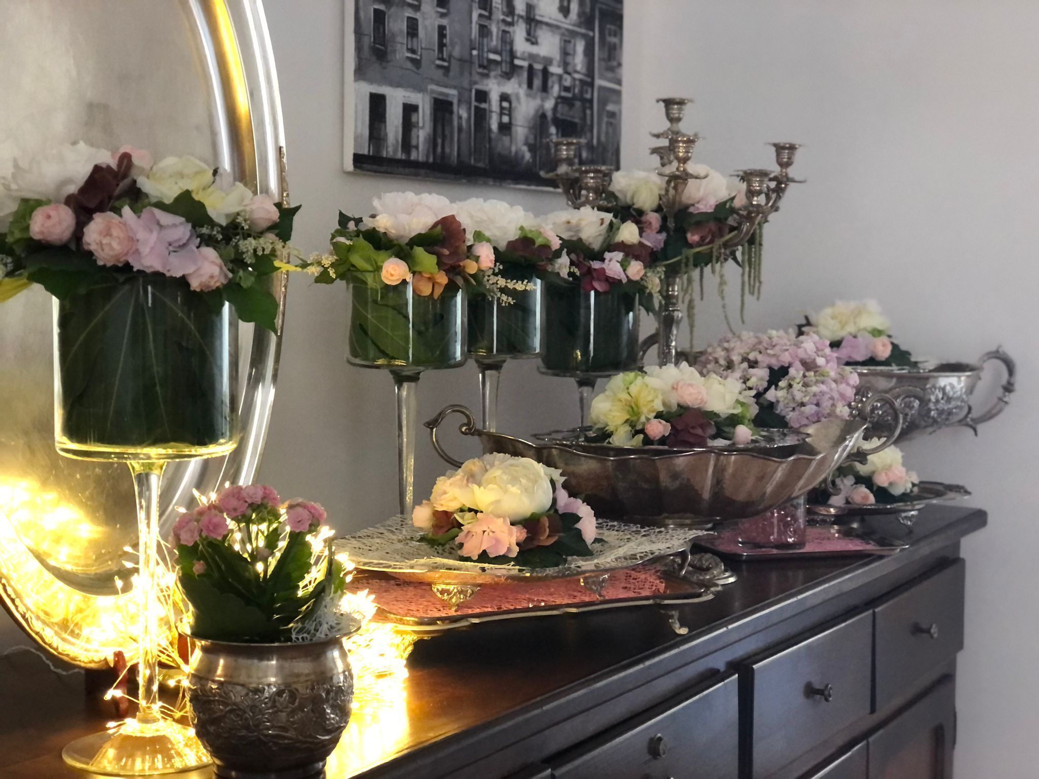 A mirror with a black and white photo, a silver candelabra, and multiple flower arrangements on a dresser.