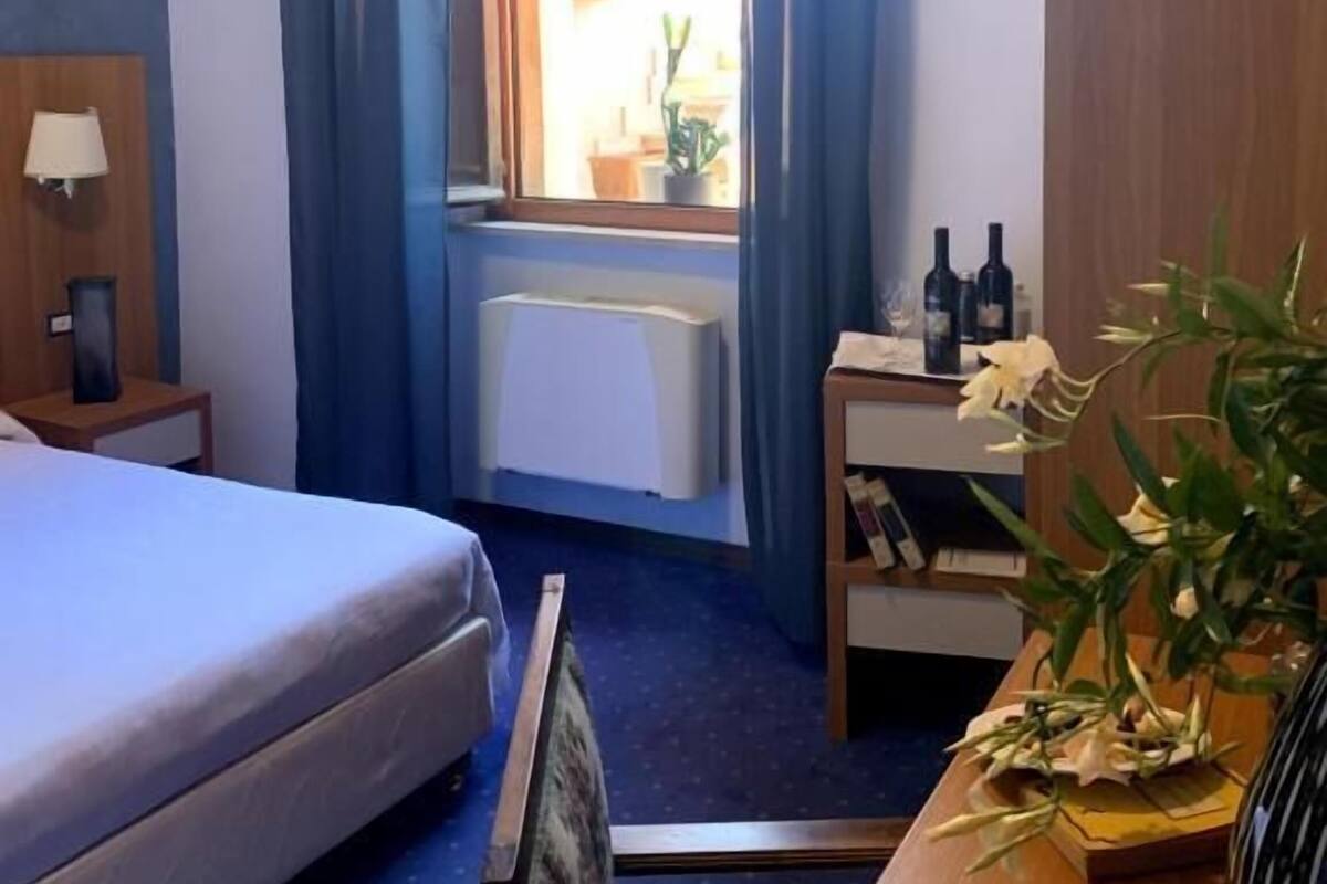 Een hotelkamer met een bed, een bureau met lamp, een raam met gordijnen, een radiator en een tafeltje met flesjes en bloemen.