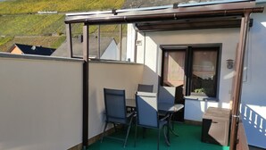 Un pequeño balcón exterior con dos sillas y una mesa, una ventana con cortinas y vistas a una colina.