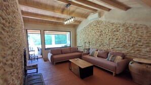 A cozy living room with a stone wall, wooden ceiling beams, a sofa, a coffee table, and a fireplace. - Rural house with garden, barbecue and swimming pool. Sea, mountains, culture, hiking! (Càlig)