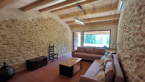 A room with stone walls, a wooden ceiling, a sofa, a table, a chair, and a window with a view. - Rural house with garden, barbecue and swimming pool. Sea, mountains, culture, hiking! (Càlig)