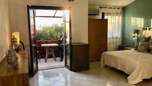 Un dormitorio con cama, un armario de madera, una mesa con lámpara y un balcón con mesa y sillas.