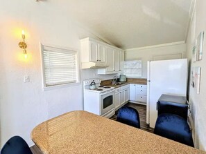 Cocina compacta con armarios blancos, estufa y refrigerador. Cuenta con un pequeño comedor con una mesa y dos sillas.