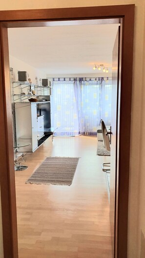 Une cuisine moderne équipée d’appareils électroménagers en acier inoxydable, un coin repas avec une table et des chaises, et une vue sur le salon, accessible par une porte, avec une grande fenêtre et des rideaux.