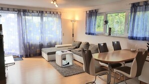 Un salon moderne avec un canapé, une table basse et une table à manger avec des chaises. On y trouve également un lampadaire et une fenêtre avec des rideaux.