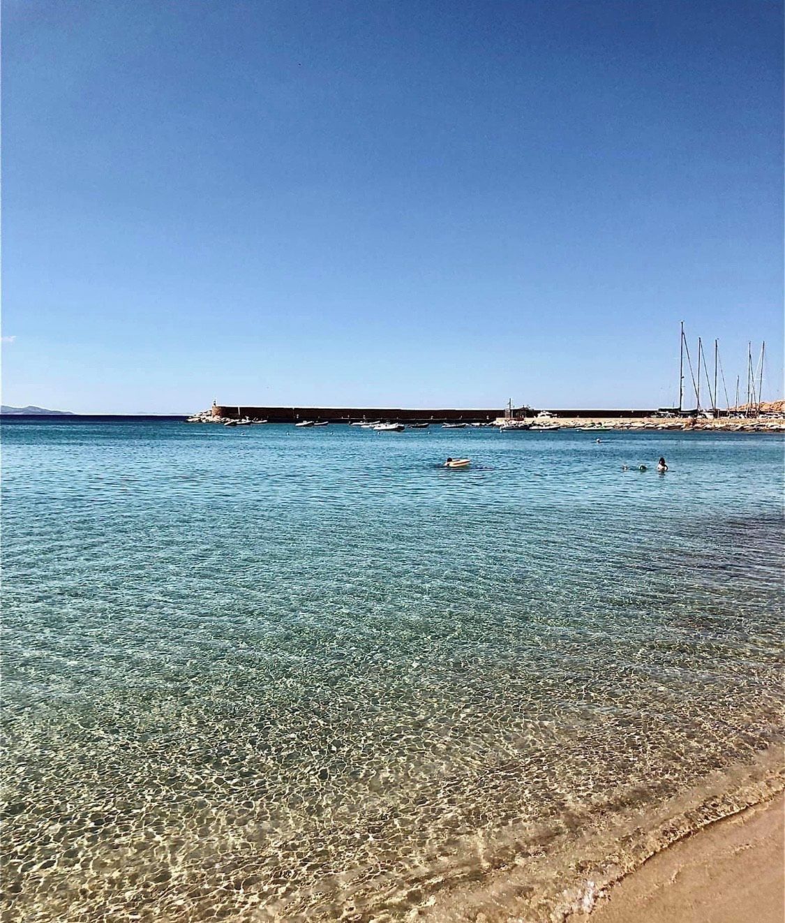 Ein sonniger Strand mit ruhigem Wasser, einem sandigen Ufer und einem entfernten Steg mit Booten.