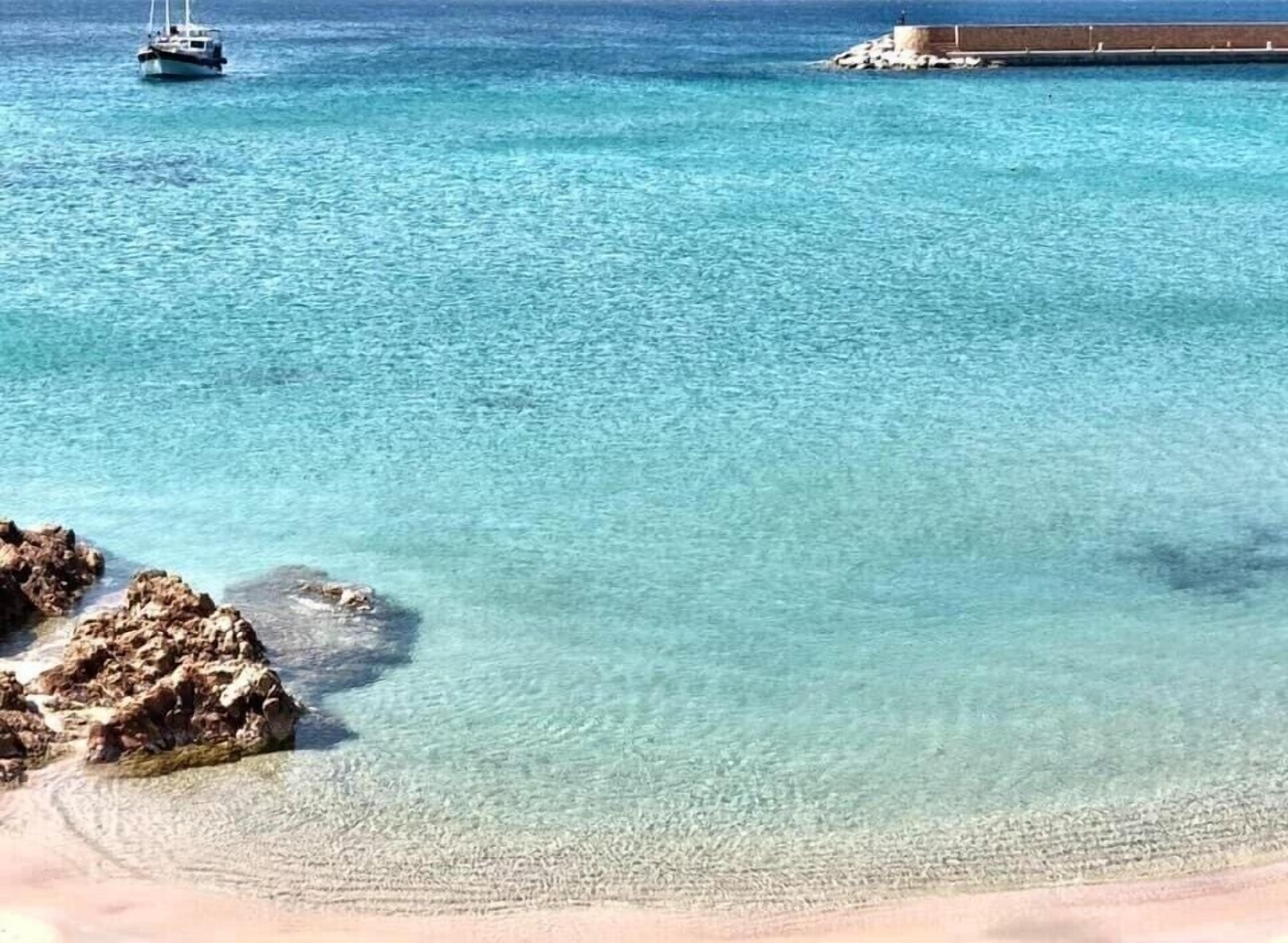 Eine Küstenansicht mit klarem türkisblauem Wasser, einem Boot, Felsen und einem Betonsteg.