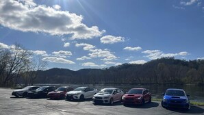 A parking lot with several cars parked near a body of water and forested hills.