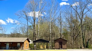 A rustic cabin with a porch, a covered picnic area, and a small shed, surrounded by tall trees and a clear sky.