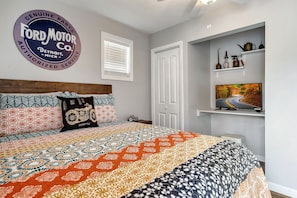 A bedroom with a patterned bedspread, a mounted Ford Motor Co. sign, a television on a shelf, and a window with blinds.