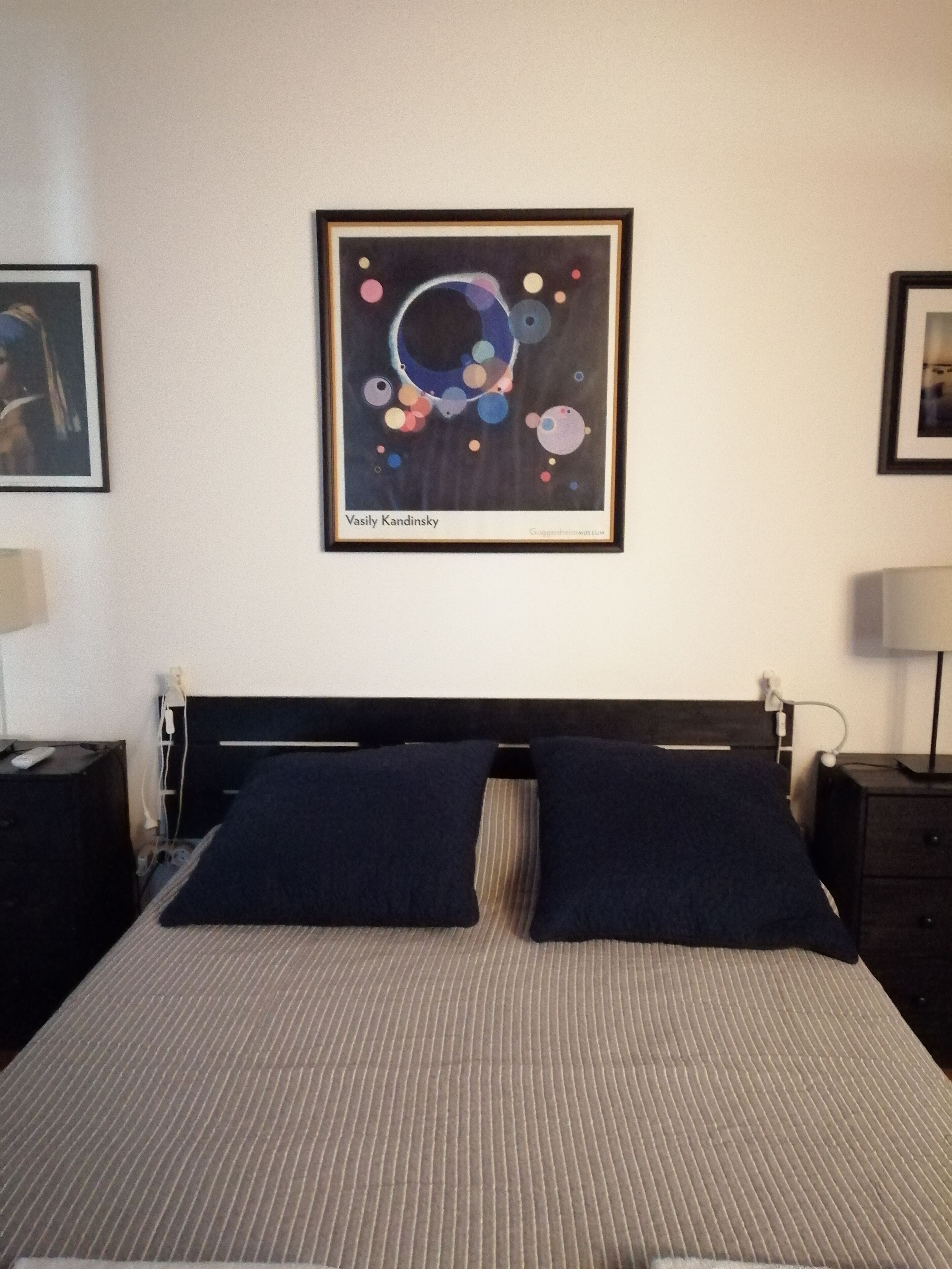 Een hotelkamer met een bed, twee kussens, een lijst met een abstract schilderij en een nachtkastje met een lamp.