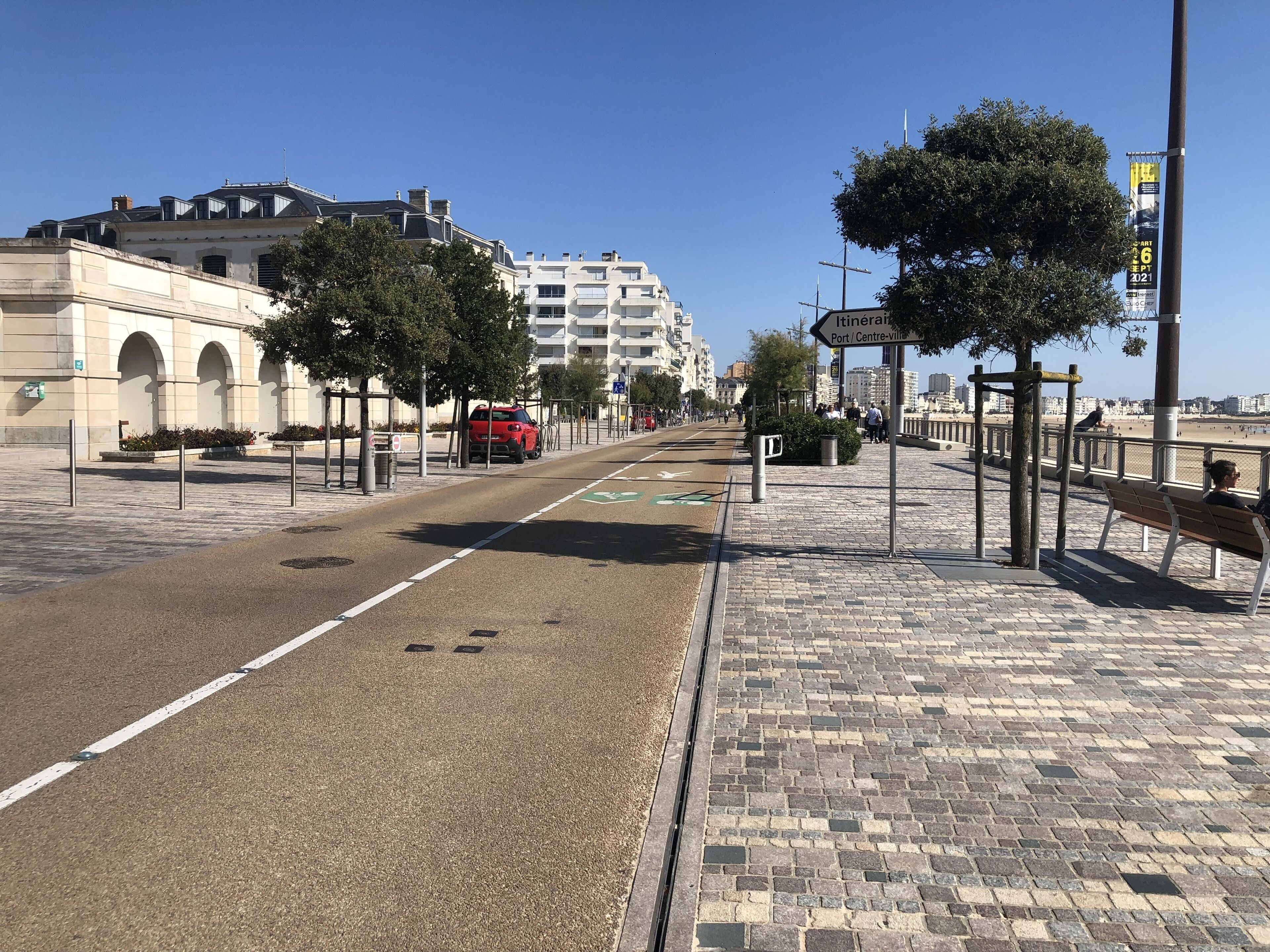 Une rue pavée avec une piste cyclable, une voiture rouge et un trottoir pour les piétons agrémenté de bancs.