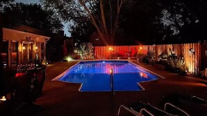 A swimming pool with a wooden deck and lounge chairs, surrounded by trees and a building with warm lighting.