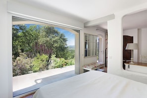 Ein Schlafzimmer mit großem Fenster, einem Bett, einem Nachttisch und einer Lampe.