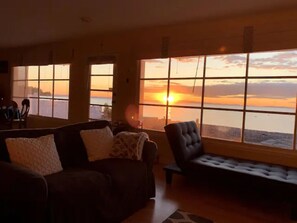 Sala de estar com uma grande janela que dá para um pôr do sol, um sofá e uma poltrona.