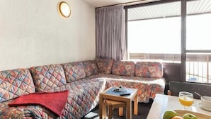 Ein gemütliches Wohnzimmer mit einer gemusterten Couch, einem kleinen Holztisch mit einem Glas Orangensaft und einem Fenster mit Vorhängen.