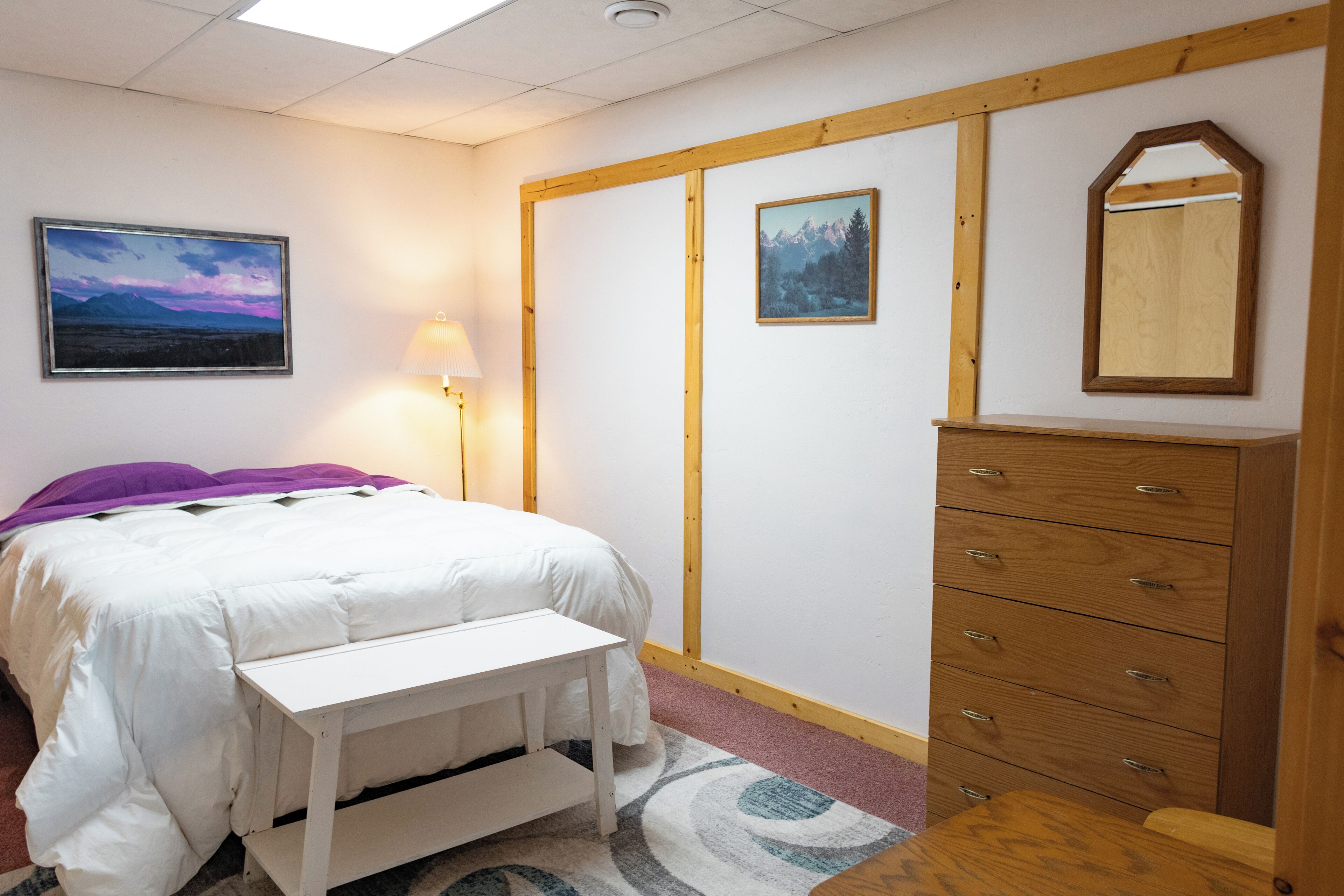 A bedroom with a bed, a bedside table, a lamp, a dresser, and two framed pictures on the wall.