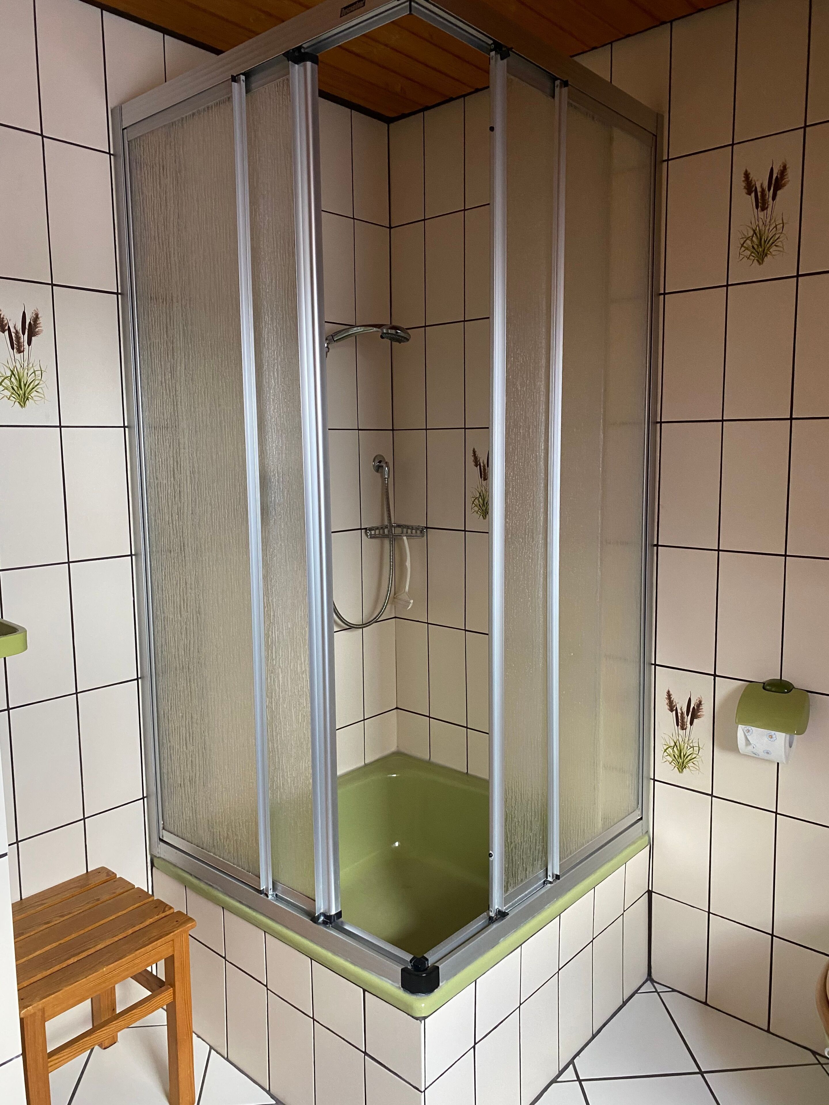 Een douchecabine met een glazen deur, een handdouche en een klein houten krukje.