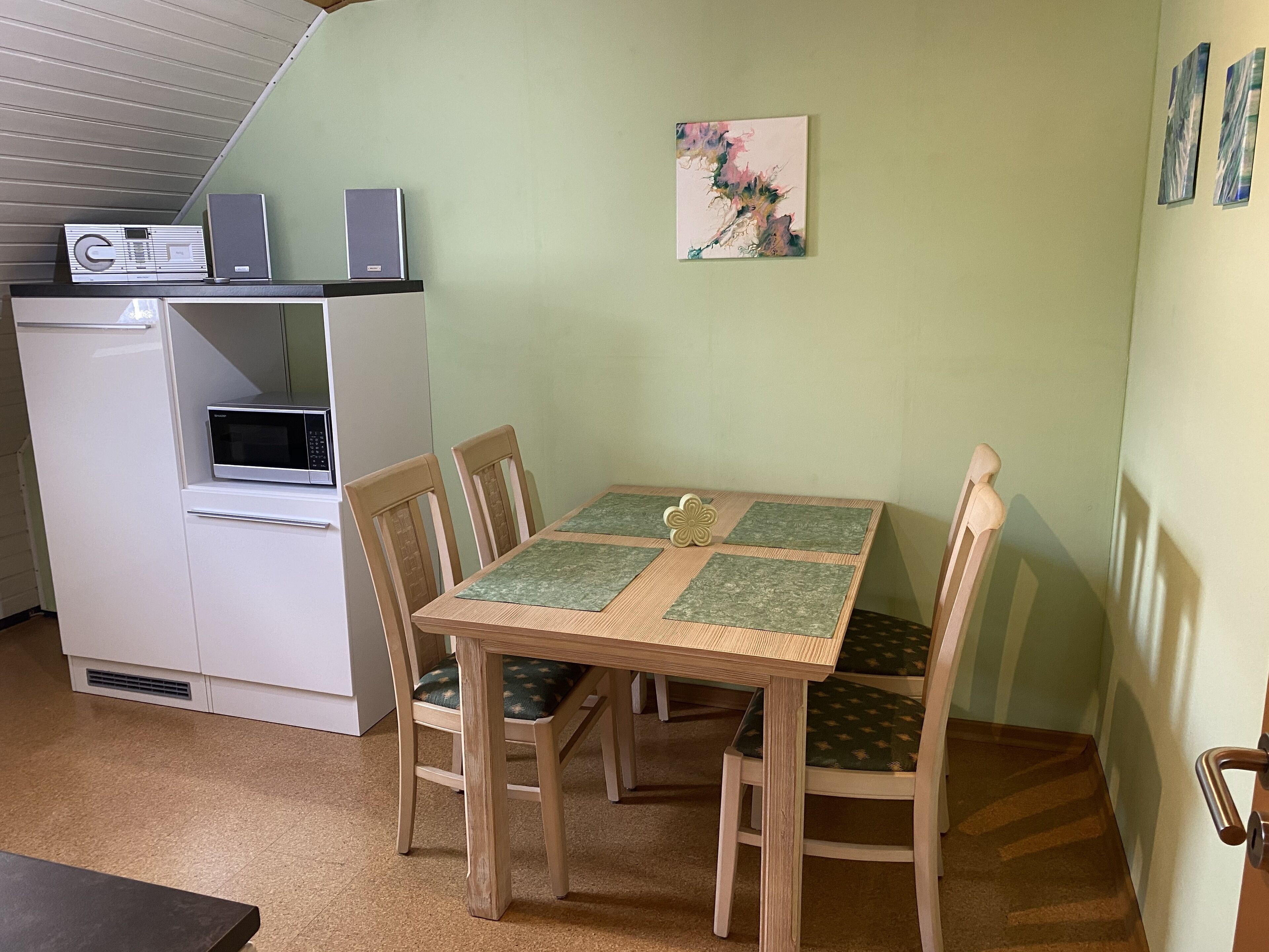 A compact room with a dining table, four chairs, a small kitchenette, and a single artwork on the wall.