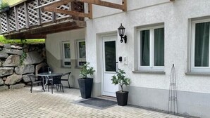 A building with a white exterior, wooden beams, a stone wall, a door, windows, and a small patio area with a table and chairs. - Holiday apartment at Ohrensbächle (Glottertal)