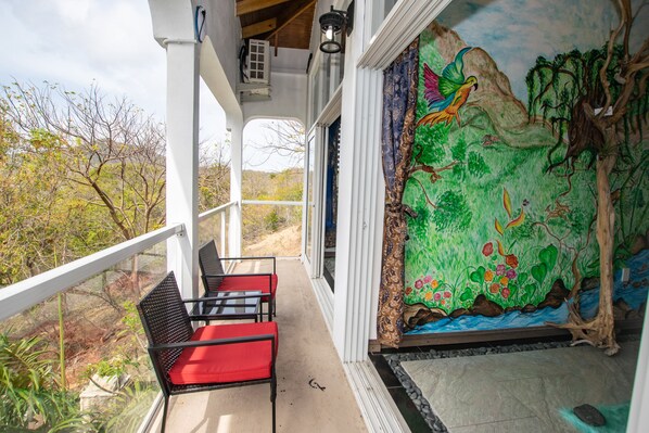 A balcony with red cushioned chairs, a mural of a tropical scene, and a view of trees and hills. - Luxurious one bedroom cottage (Carriacou and Petite Martinique)