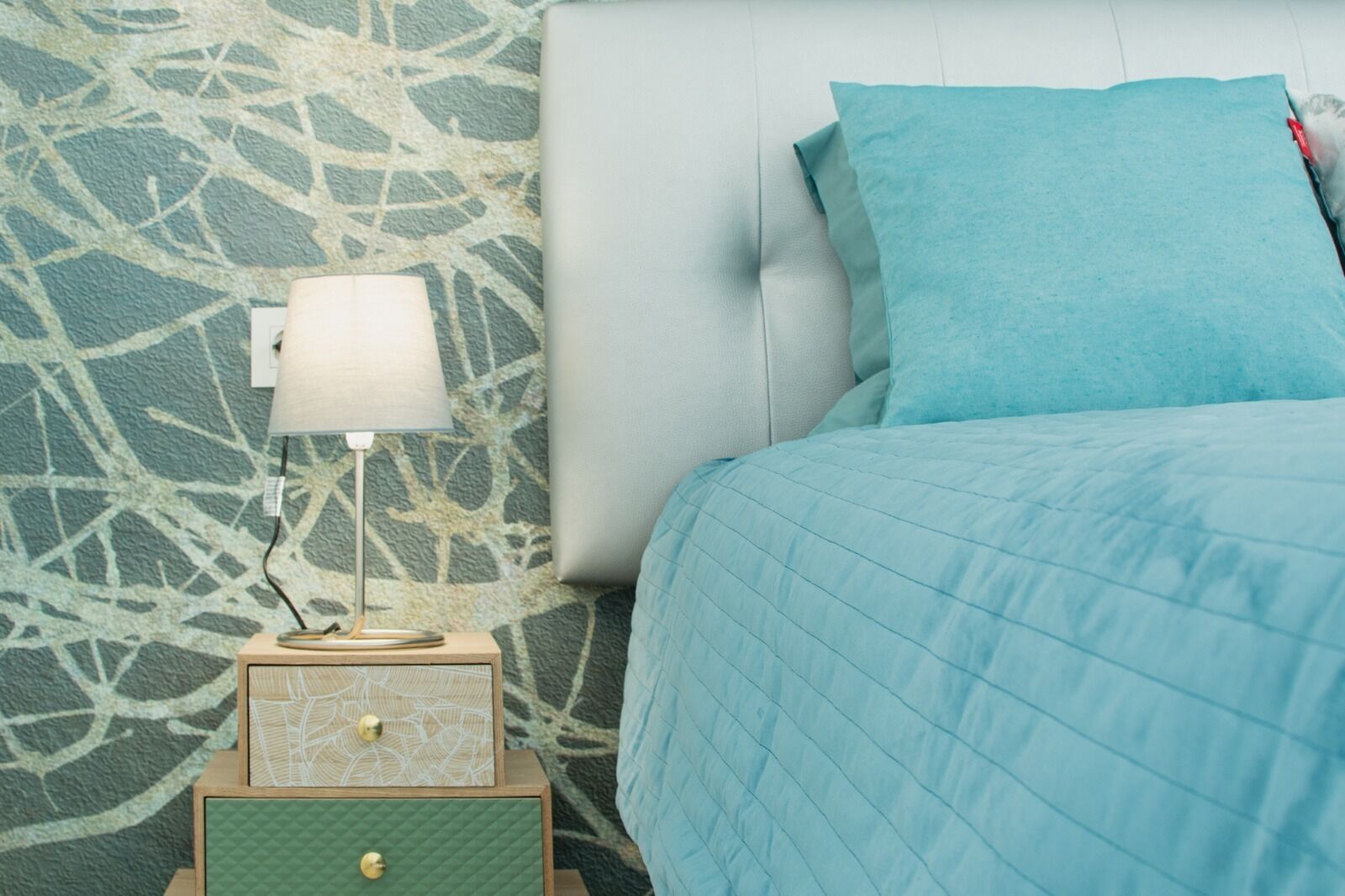 Ein Bett mit blauer Steude, ein Nachttisch mit Lampe und eine Wand mit Muster.