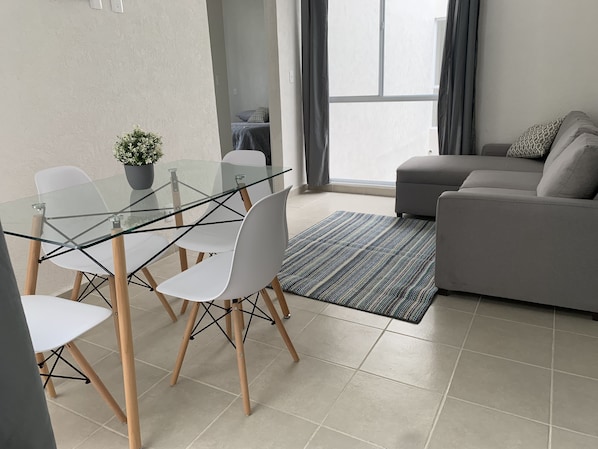 Un salon moderne avec une table à manger en verre, des chaises blanches, un canapé gris et un tapis à motifs.