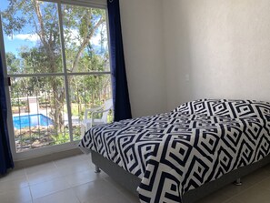 Une chambre avec un lit, une fenêtre donnant sur une piscine et des arbres, et un mur blanc.