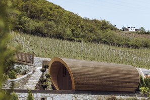 A wooden barrel-shaped structure with a stone staircase leading up to it, surrounded by a vineyard and a hillside with a house in the background. - Weingut und Genusshotel Spitalerhof (Chiusa)