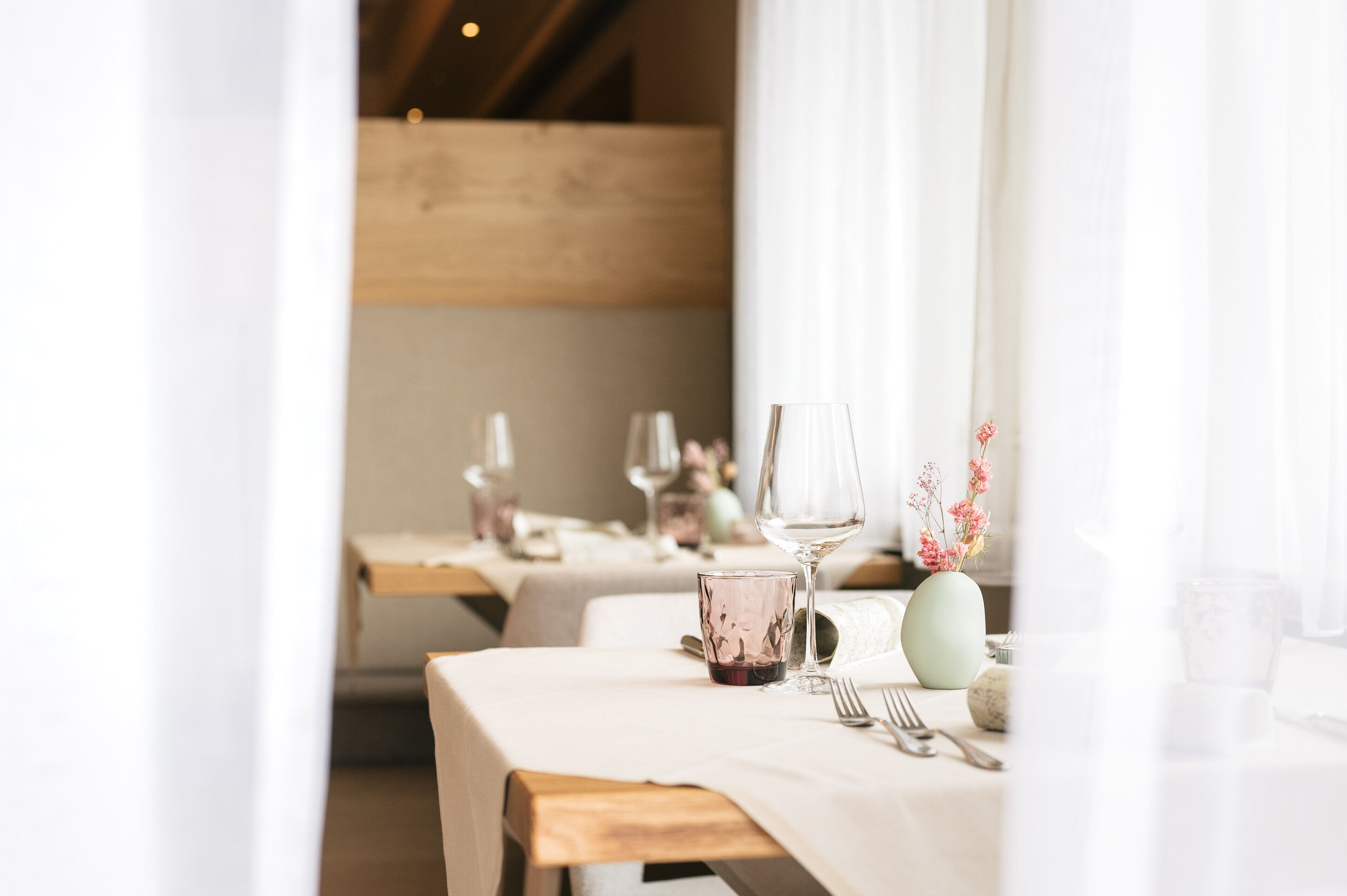 A dining area with tables set for two, featuring white tablecloths, wine glasses, and a vase with flowers.