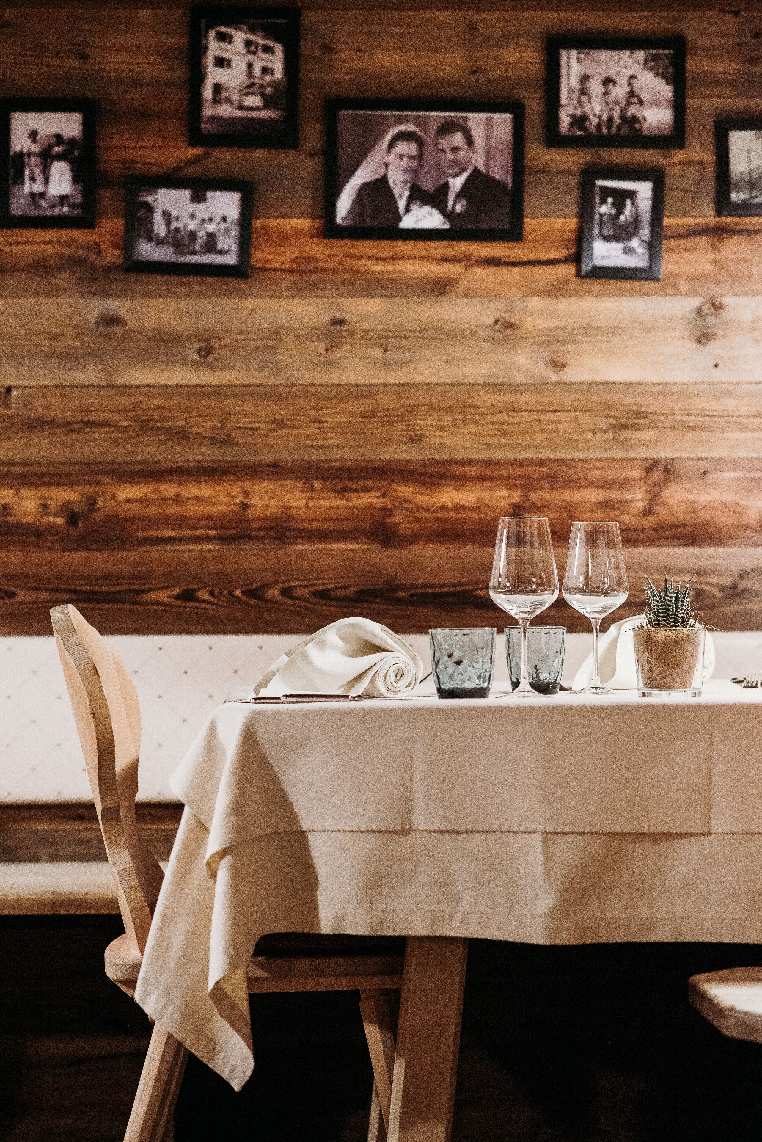 A wooden wall with framed photos, a table set with wine glasses and a potted plant.
