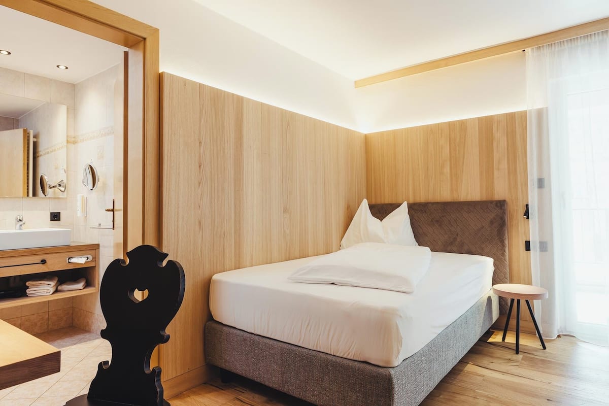 Une chambre d’hôtel moderne avec un grand lit, une tête de lit en bois, une table de chevet, un miroir et un petit tabouret.