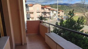 Un balcón con suelo de baldosas, barandilla metálica y vistas a edificios residenciales y vegetación al fondo.