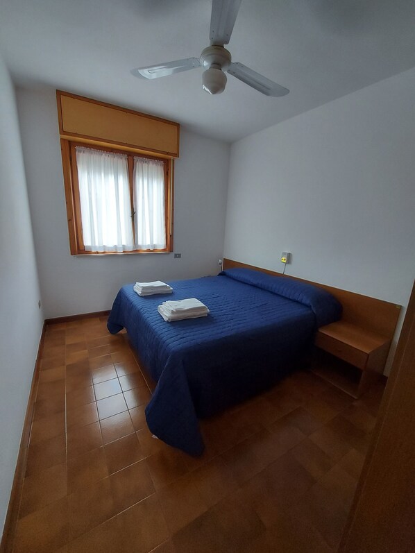 Quarto pequeno e limpo, com uma cama de solteiro, ventilador de teto e janela com cortinas.
