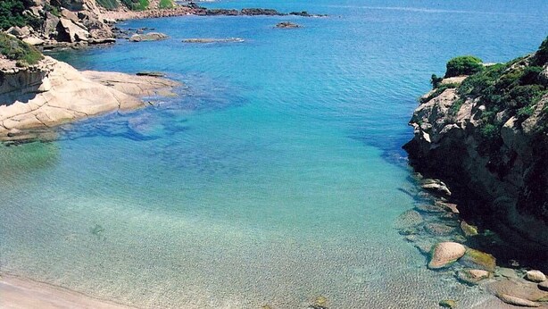 Una playa costera con aguas cristalinas, costas rocosas y montañas al fondo.