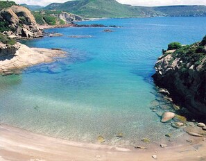 Uma praia costeira com águas claras, costas rochosas e montanhas ao fundo.