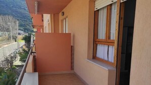 Un balcón con suelo de baldosas, una ventana con persianas y vistas a una calle y colinas.