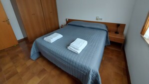 Una cama bien hecha con un edredón azul, dos toallas blancas dobladas, un armario de madera, una mesita de noche con una lámpara y un enchufe de pared.