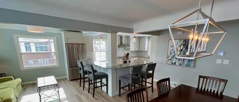 A modern kitchen with a central island, dining area with chairs, and a large window.